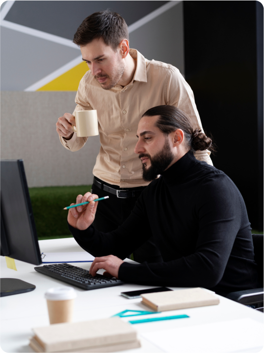 Two professionals collaborating at a desk, one holding a coffee cup while discussing ideas in front of a computer.