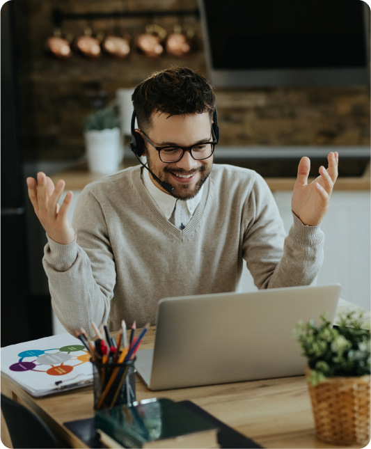 Cheerful man with glasses and a headset, engaging in a video call while smiling and gesturing at his laptop.